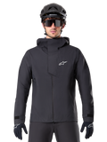 Chaqueta impermeable Alpinestars A-Dura Elite, negra, chaqueta de ciclismo de montaña, diseño de cuello alto con capucha, cremallera frontal con solapa protectora en el pecho y logotipo blanco, usada con casco negro y guantes de ciclismo, protección técnica contra la intemperie para rodar por senderos