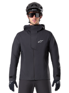 Chaqueta impermeable Alpinestars A-Dura Elite, negra, chaqueta de ciclismo de montaña, diseño de cuello alto con capucha, cremallera frontal con solapa protectora en el pecho y logotipo blanco, usada con casco negro y guantes de ciclismo, protección técnica contra la intemperie para rodar por senderos
