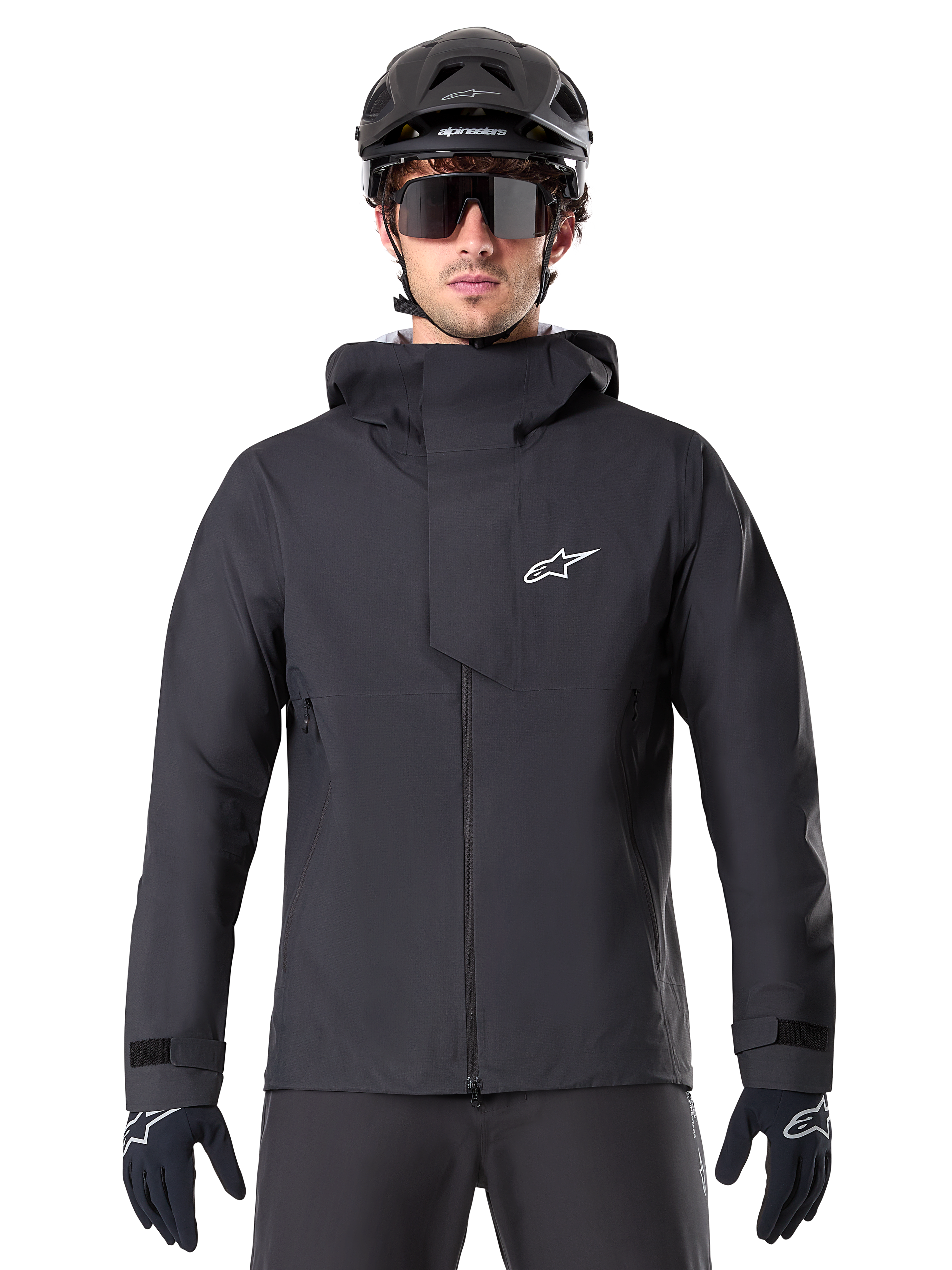 Chaqueta impermeable Alpinestars A-Dura Elite, negra, chaqueta de ciclismo de montaña, diseño de cuello alto con capucha, cremallera frontal con solapa protectora en el pecho y logotipo blanco, usada con casco negro y guantes de ciclismo, protección técnica contra la intemperie para rodar por senderos