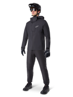 Chaqueta impermeable Alpinestars A-Dura Elite en color negro, usada por un ciclista con pantalones de MTB, guantes y casco negros a juego. Presenta un diseño técnico impermeable con el logotipo Astars blanco en el pecho y un cuello protector alto.