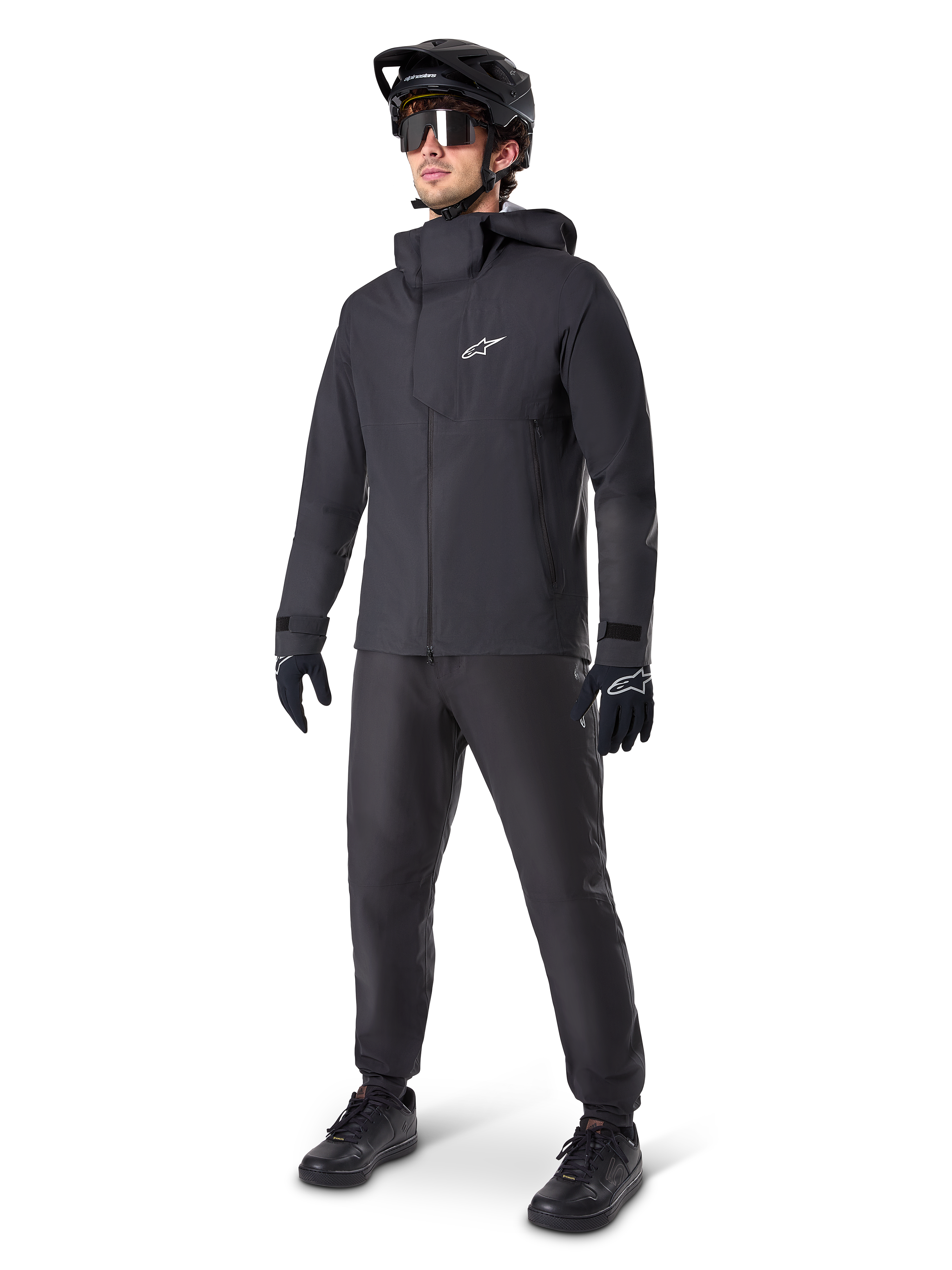 Chaqueta impermeable Alpinestars A-Dura Elite en color negro, usada por un ciclista con pantalones de MTB, guantes y casco negros a juego. Presenta un diseño técnico impermeable con el logotipo Astars blanco en el pecho y un cuello protector alto.