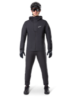 Chaqueta impermeable Alpinestars A-Dura Elite, negra, combinada con pantalones y guantes de ciclismo a juego, casco negro y gafas de sol, con el logotipo de Astars en blanco en el pecho, diseño técnico de alto rendimiento para ciclismo de montaña