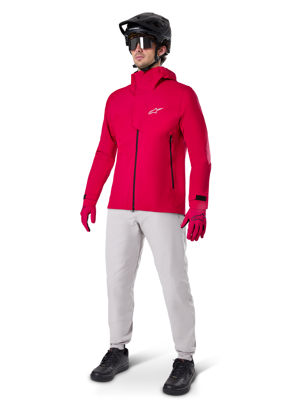 Ciclista masculino con la chaqueta impermeable Alpinestars A-Dura Elite en rojo flúor, combinada con pantalones técnicos gris claro, guantes rojos, casco de MTB negro con gafas y zapatillas negras, equipamiento completo de alto rendimiento para ciclismo de montaña