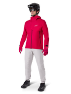 Ciclista masculino con la chaqueta impermeable Alpinestars A-Dura Elite en rojo flúor, combinada con pantalones técnicos gris claro, guantes rojos, casco de MTB negro con gafas y zapatillas negras, equipamiento completo de alto rendimiento para ciclismo de montaña