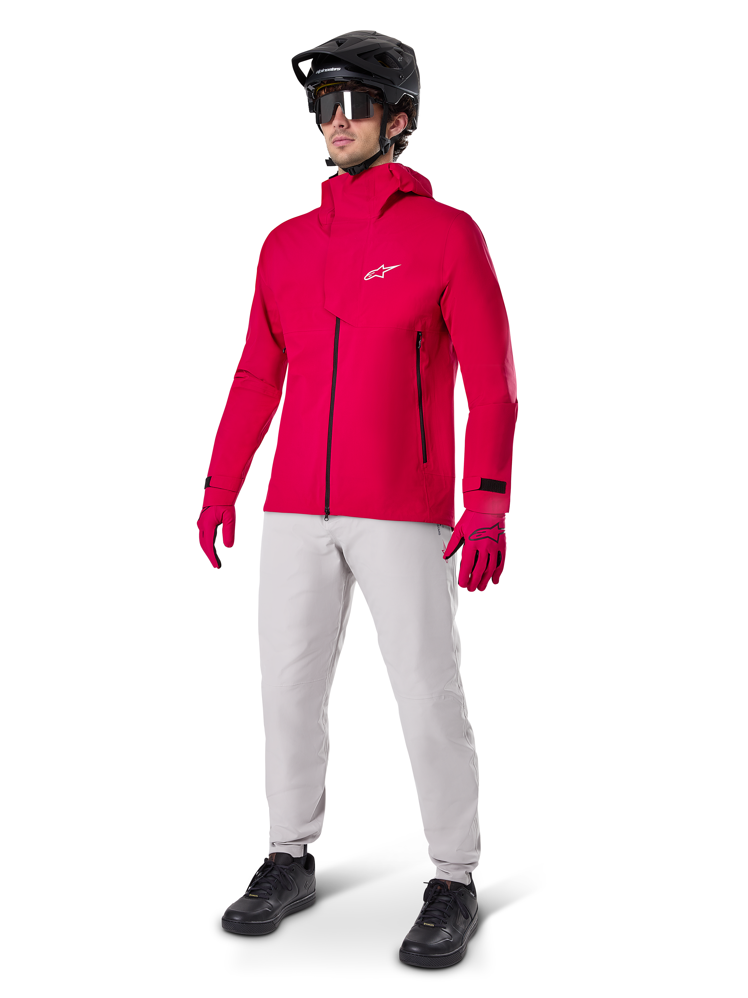 Ciclista masculino con la chaqueta impermeable Alpinestars A-Dura Elite en rojo flúor, combinada con pantalones técnicos gris claro, guantes rojos, casco de MTB negro con gafas y zapatillas negras, equipamiento completo de alto rendimiento para ciclismo de montaña