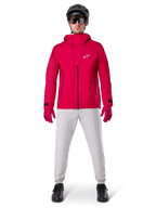 Chaqueta impermeable Alpinestars A-Dura Elite, Rojo Fluo, chaqueta técnica de montaña usada con un casco negro de MTB, gafas de sol, guantes rojos a juego y pantalones gris claro, vista de cuerpo completo del equipamiento de ciclismo para todo tipo de clima