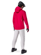 Alpinestars A-Dura Elite, chaqueta impermeable, Rojo Fluo, vista trasera de tres cuartos, combinada con pantalones gris claro y casco de MTB negro, cuenta con capucha y detalles reflectantes, chaqueta de lluvia de alto rendimiento para ciclismo de montaña