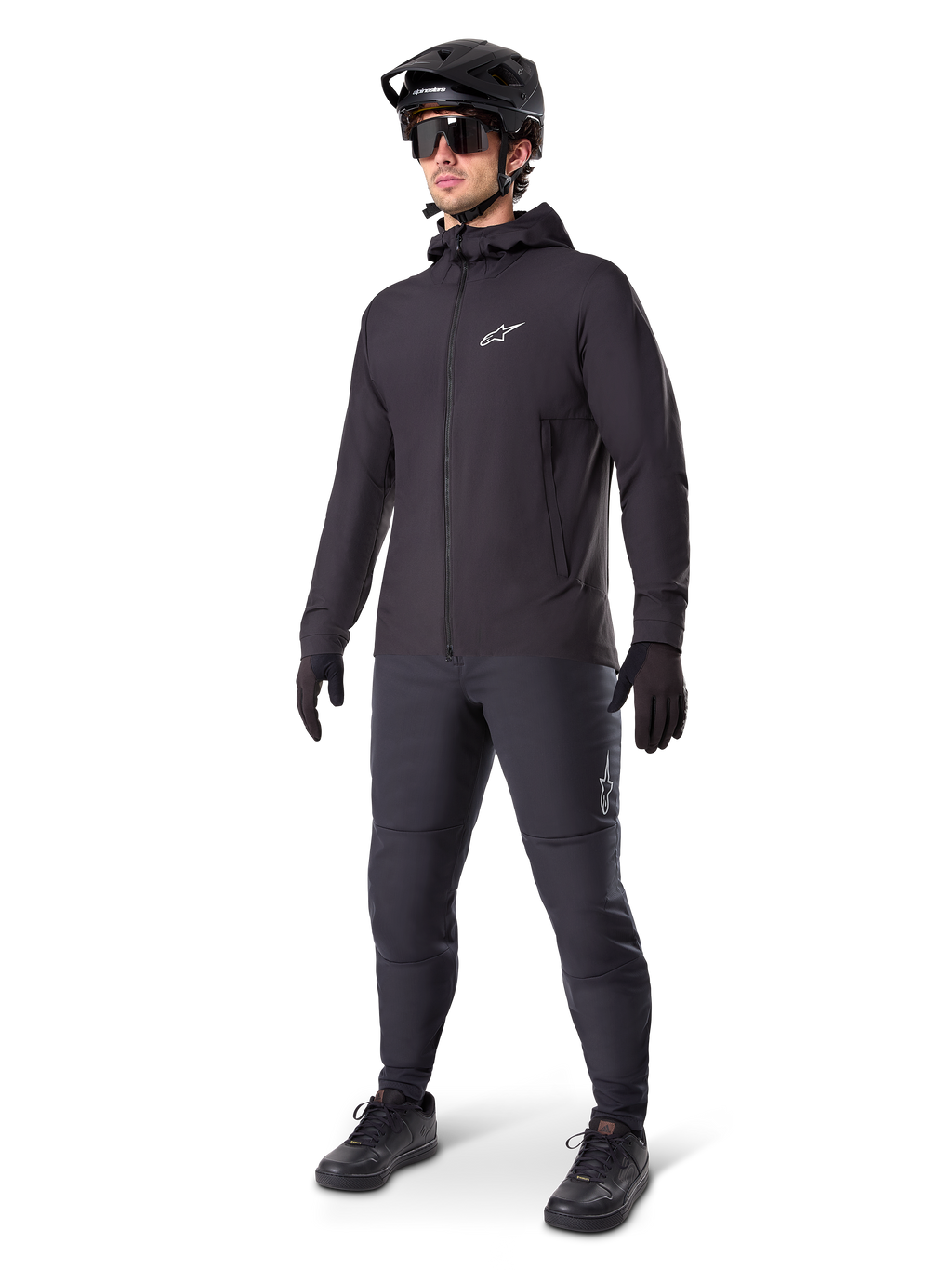 Chaqueta térmica Alpinestars A-Dura en color negro, usada por un ciclista con equipamiento completo de mountain bike, incluyendo casco negro, gafas de sol y pantalones de ciclismo; chaqueta ligera con capucha y aislamiento para un alto rendimiento en ciclismo en climas fríos.