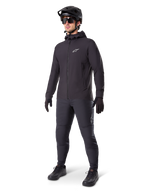 Chaqueta térmica Alpinestars A-Dura en color negro, usada por un ciclista con equipamiento completo de mountain bike, incluyendo casco negro, gafas de sol y pantalones de ciclismo; chaqueta ligera con capucha y aislamiento para un alto rendimiento en ciclismo en climas fríos.