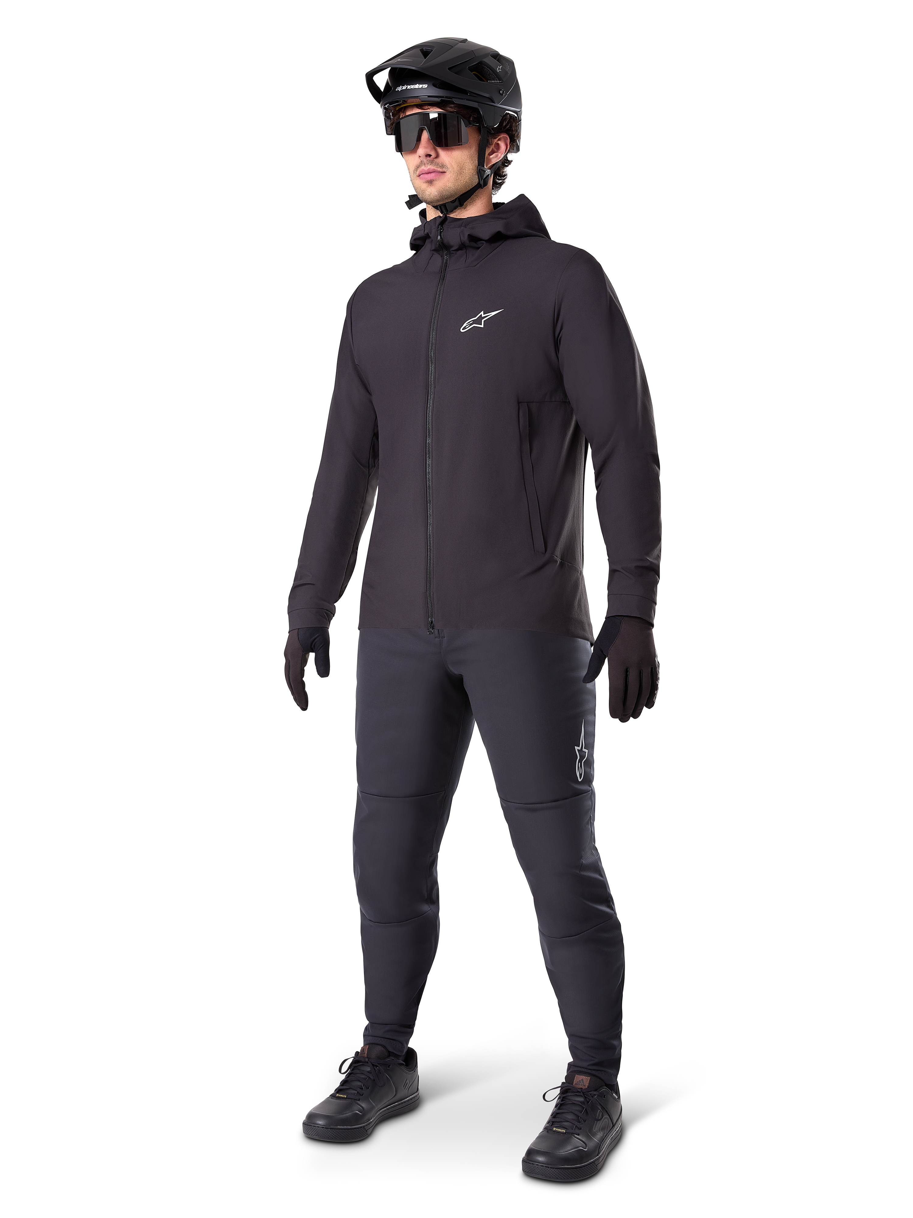 Chaqueta térmica Alpinestars A-Dura en color negro, usada por un ciclista con equipamiento completo de mountain bike, incluyendo casco negro, gafas de sol y pantalones de ciclismo; chaqueta ligera con capucha y aislamiento para un alto rendimiento en ciclismo en climas fríos.