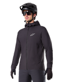 Alpinestars A-Dura Thermal Jacket, chaqueta de ciclismo negra, diseño con capucha y cremallera frontal, logotipo de estrella blanca en el pecho, usada con casco de bicicleta de montaña y guantes negros, equipamiento deportivo de alto rendimiento para rutas en climas fríos