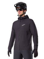 Alpinestars A-Dura Thermal Jacket, chaqueta de ciclismo negra, diseño con capucha y cremallera frontal, logotipo de estrella blanca en el pecho, usada con casco de bicicleta de montaña y guantes negros, equipamiento deportivo de alto rendimiento para rutas en climas fríos