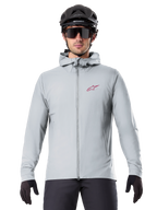 Chaqueta térmica Alpinestars A-Dura en color Titanium, chaqueta de ciclismo con capucha en gris claro con un logotipo de estrella roja en el pecho, usada por un ciclista con casco de MTB negro, gafas de sol y guantes, equipamiento aislante para ciclismo de montaña