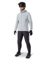 Chaqueta térmica Alpinestars A-Dura en Titanium, chaqueta de ciclismo con capucha en gris claro con logotipo rojo de Alpinestars en el pecho, usada por un hombre con casco de MTB negro, gafas de sol, pantalones gris oscuro y guantes negros, equipamiento profesional de mountain bike