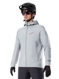 Alpinestars A-Dura Thermal Jacket, chaqueta de ciclismo gris claro con aislamiento, diseño con capucha y un pequeño logotipo rojo, usada con casco y guantes negros, ropa técnica de mountain bike para climas fríos