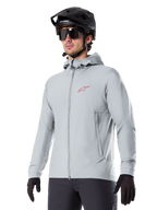 Alpinestars A-Dura Thermal Jacket, chaqueta de ciclismo gris claro con aislamiento, diseño con capucha y un pequeño logotipo rojo, usada con casco y guantes negros, ropa técnica de mountain bike para climas fríos