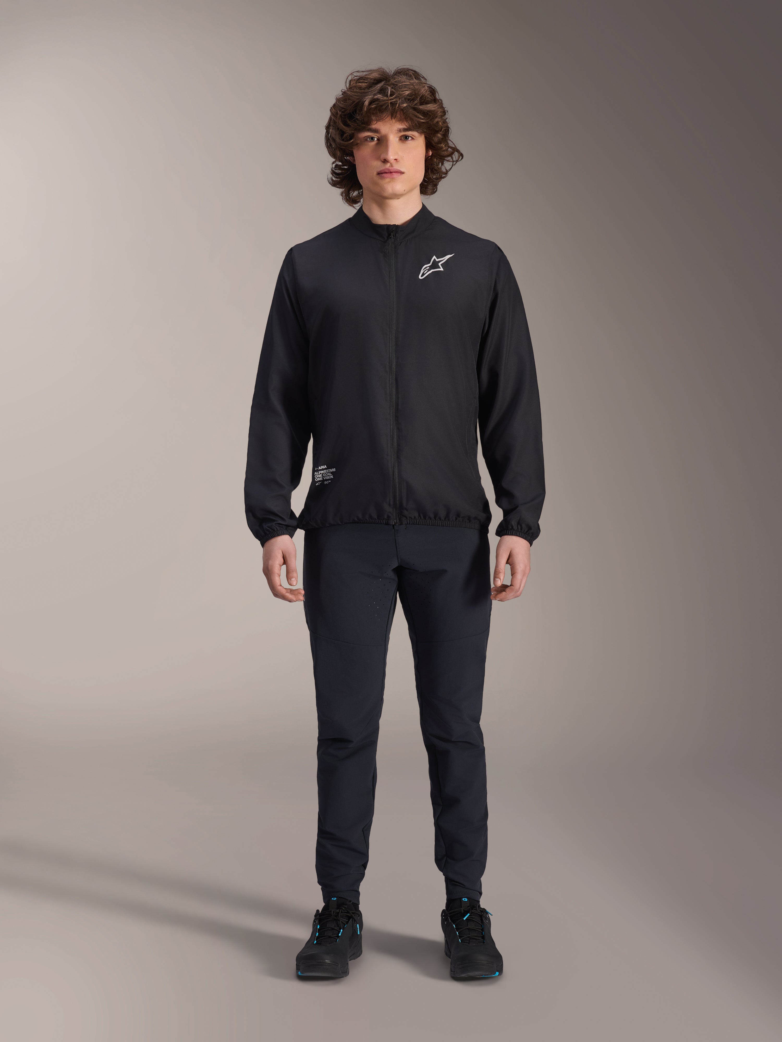 Modelo con la chaqueta cortavientos Alpinestars A-Aria en color negro, con cremallera frontal completa, cuello simulado y logotipo Astars blanco en el pecho, combinada con pantalones técnicos negros y calzado negro, ropa de ciclismo de rendimiento para ciclismo de montaña y uso al aire libre.