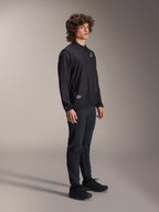 Chaqueta Cortavientos Alpinestars A-Aria, negra, chaqueta de ciclismo ligera y resistente al viento lucida por un modelo con pelo rizado, con un logotipo blanco de Astars en el pecho y marca en la parte inferior derecha, combinada con pantalones y calzado negros, ajuste de rendimiento para mountain bike y gravel