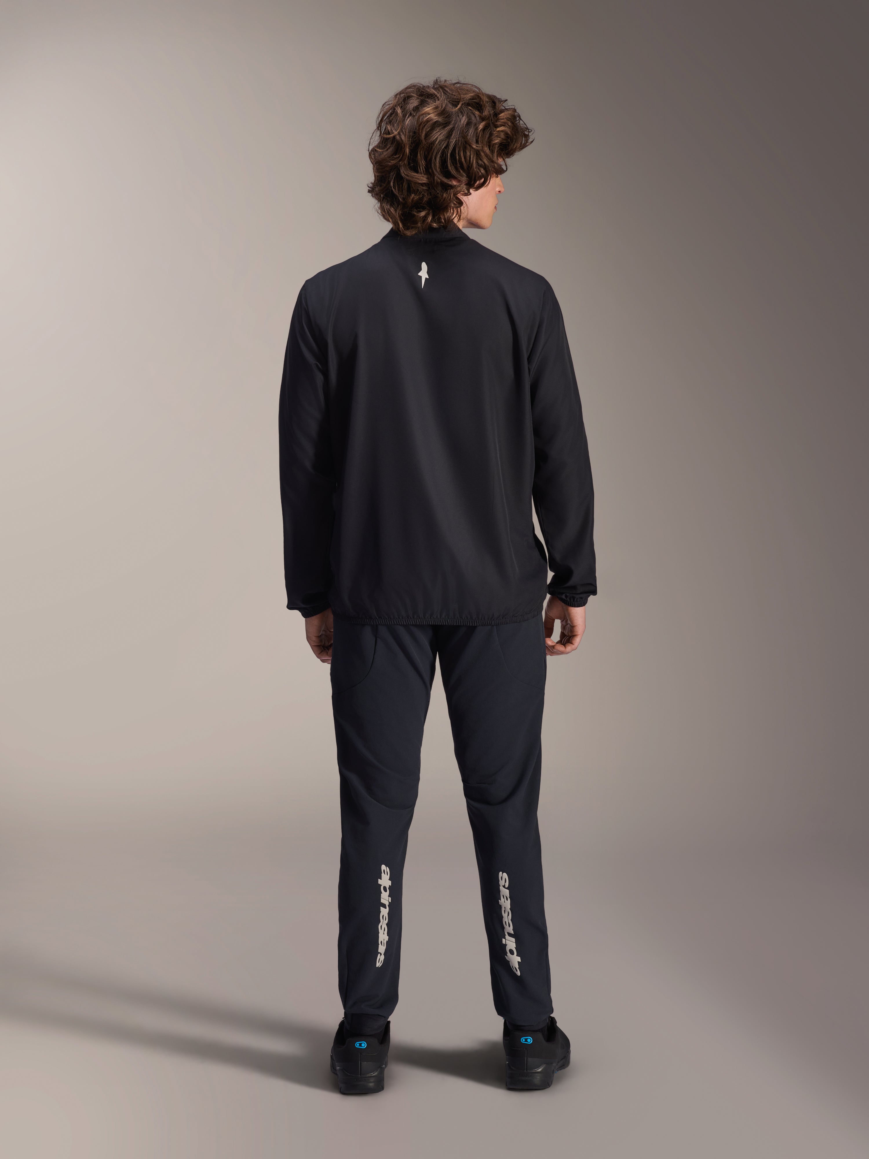 Chaqueta cortavientos Alpinestars A-Aria, negra, vista trasera, chaqueta de ciclismo ligera y resistente al viento con un pequeño logotipo blanco en la parte superior de la espalda, combinada con pantalones negros a juego con la marca Alpinestars vertical en las pantorrillas, equipamiento de alto rendimiento para bicicleta de montaña