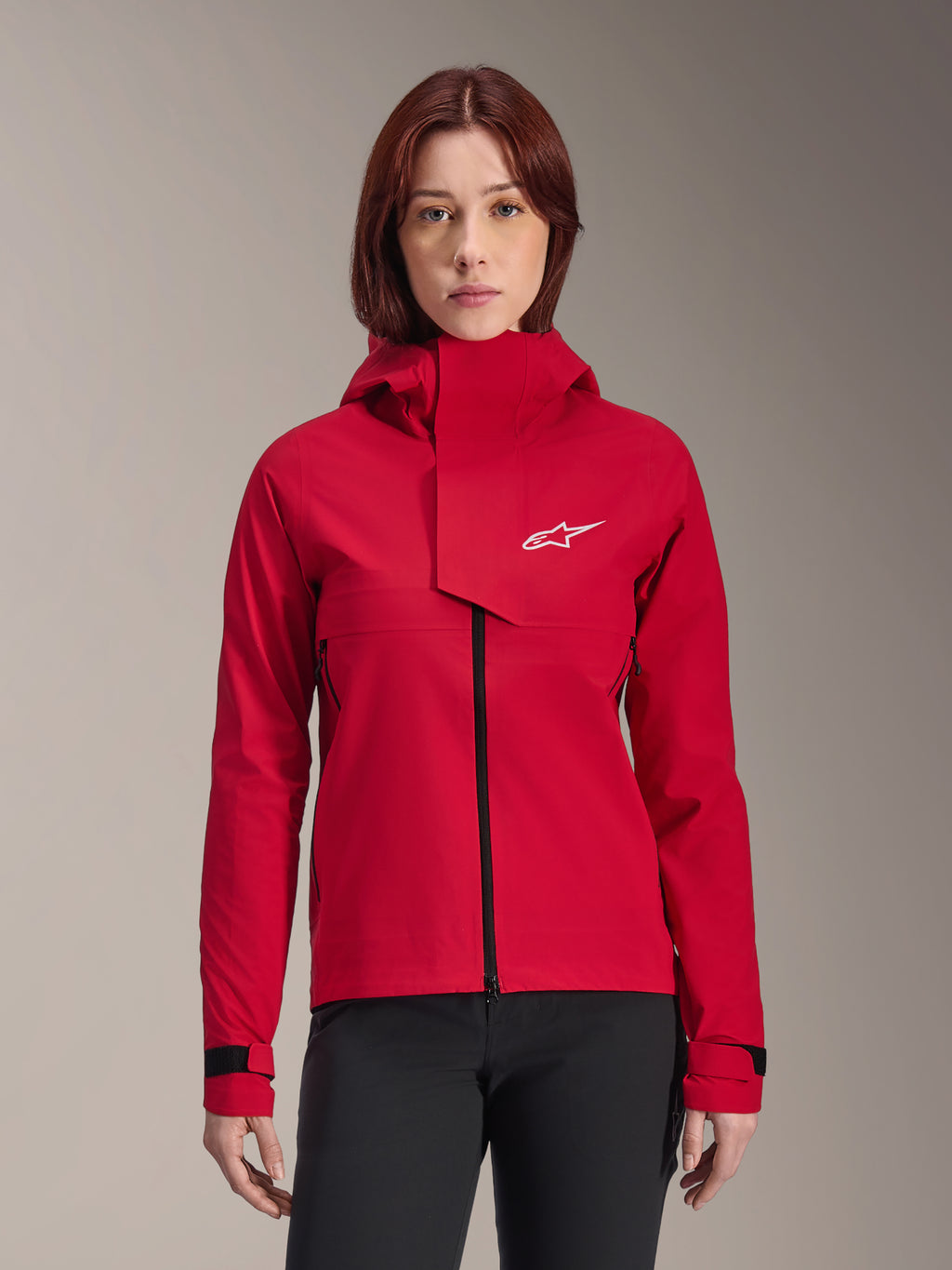 Alpinestars Stella A-Dura Elite Waterproof Jacket, chaqueta de mountain bike para mujer, rojo fluo, diseño con capucha y solapa frontal asimétrica, logotipo blanco de Alpinestars en el pecho, puños negros ajustables, lucida por una modelo con pantalones negros