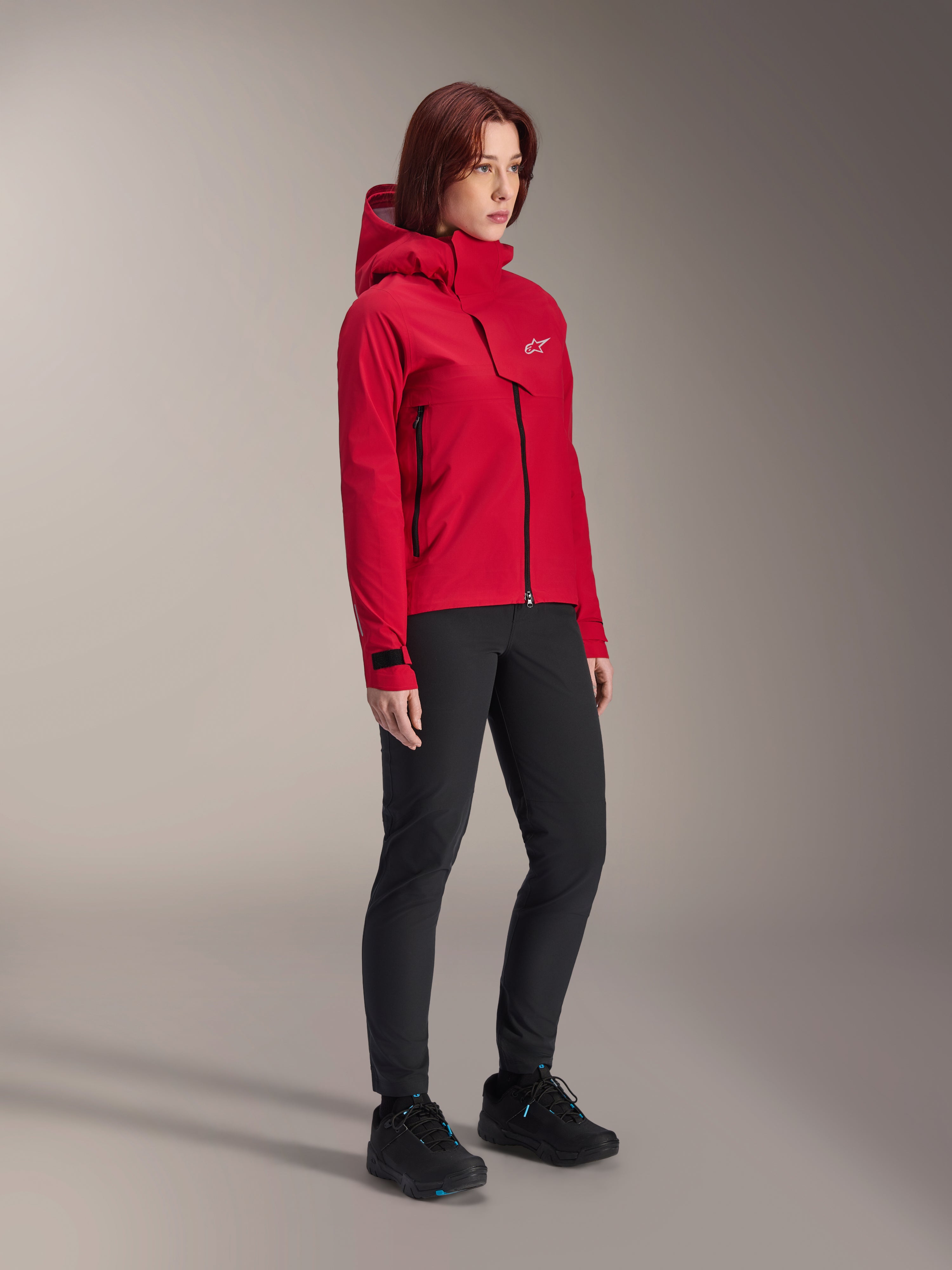 Chaqueta impermeable Alpinestars Stella A-Dura Elite para mujer, rojo flúor, chaqueta con capucha para ciclismo de montaña con el logotipo blanco de Astars en la solapa del pecho, combinada con pantalones negros y zapatillas de montaña negras, equipamiento técnico de alto rendimiento para trail