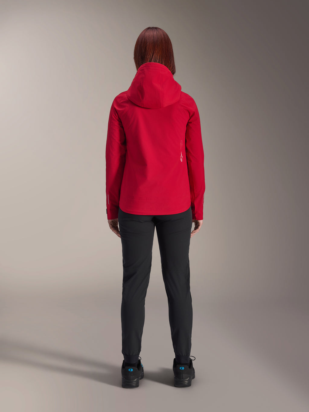 Vista posterior de una mujer con la chaqueta impermeable Alpinestars Stella A-Dura Elite en Rojo Fluo, combinada con pantalones y calzado negros, con capucha protectora y dobladillo trasero más largo para ciclismo de montaña
