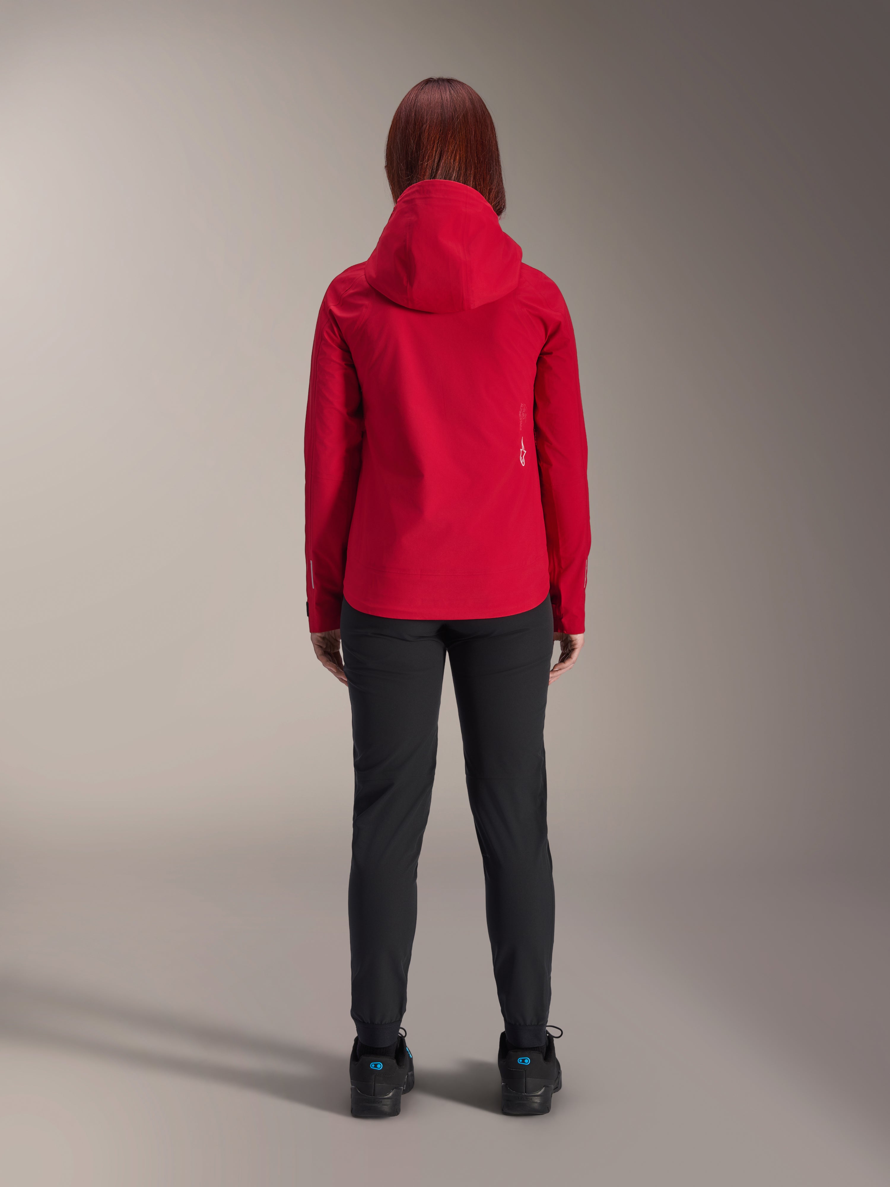 Vista posterior de una mujer con la chaqueta impermeable Alpinestars Stella A-Dura Elite en Rojo Fluo, combinada con pantalones y calzado negros, con capucha protectora y dobladillo trasero más largo para ciclismo de montaña