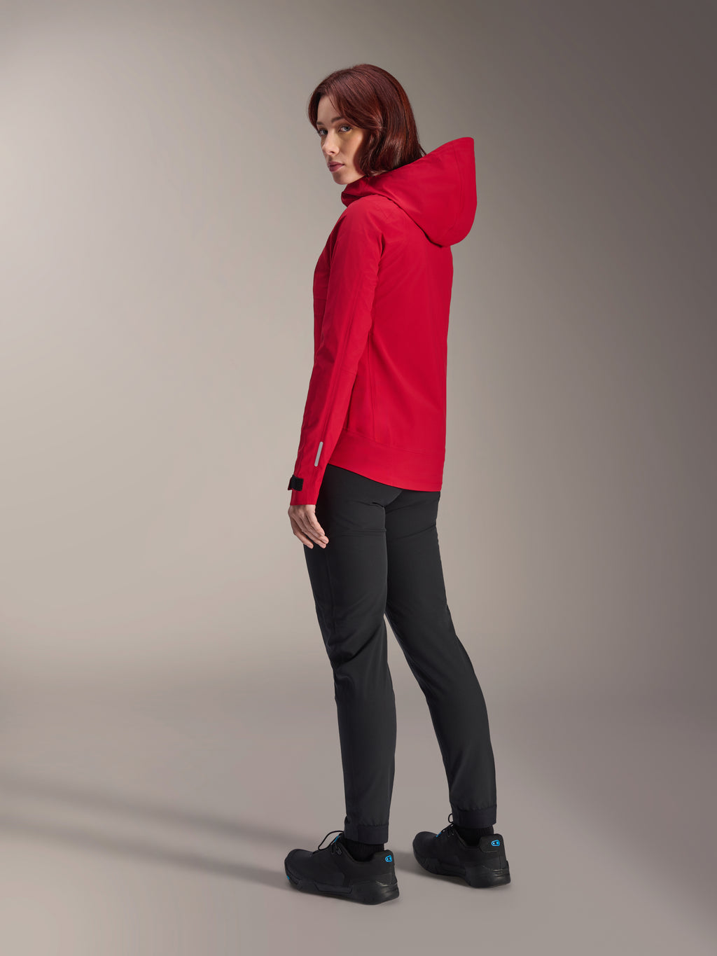 Mujer vistiendo la chaqueta impermeable Alpinestars Stella A-Dura Elite en color Red Fluo, combinada con pantalones técnicos de ciclismo negros y calzado negro, vista trasera de tres cuartos que muestra la capucha ajustable y el dobladillo trasero más largo, ropa de alto rendimiento para bicicleta de montaña.