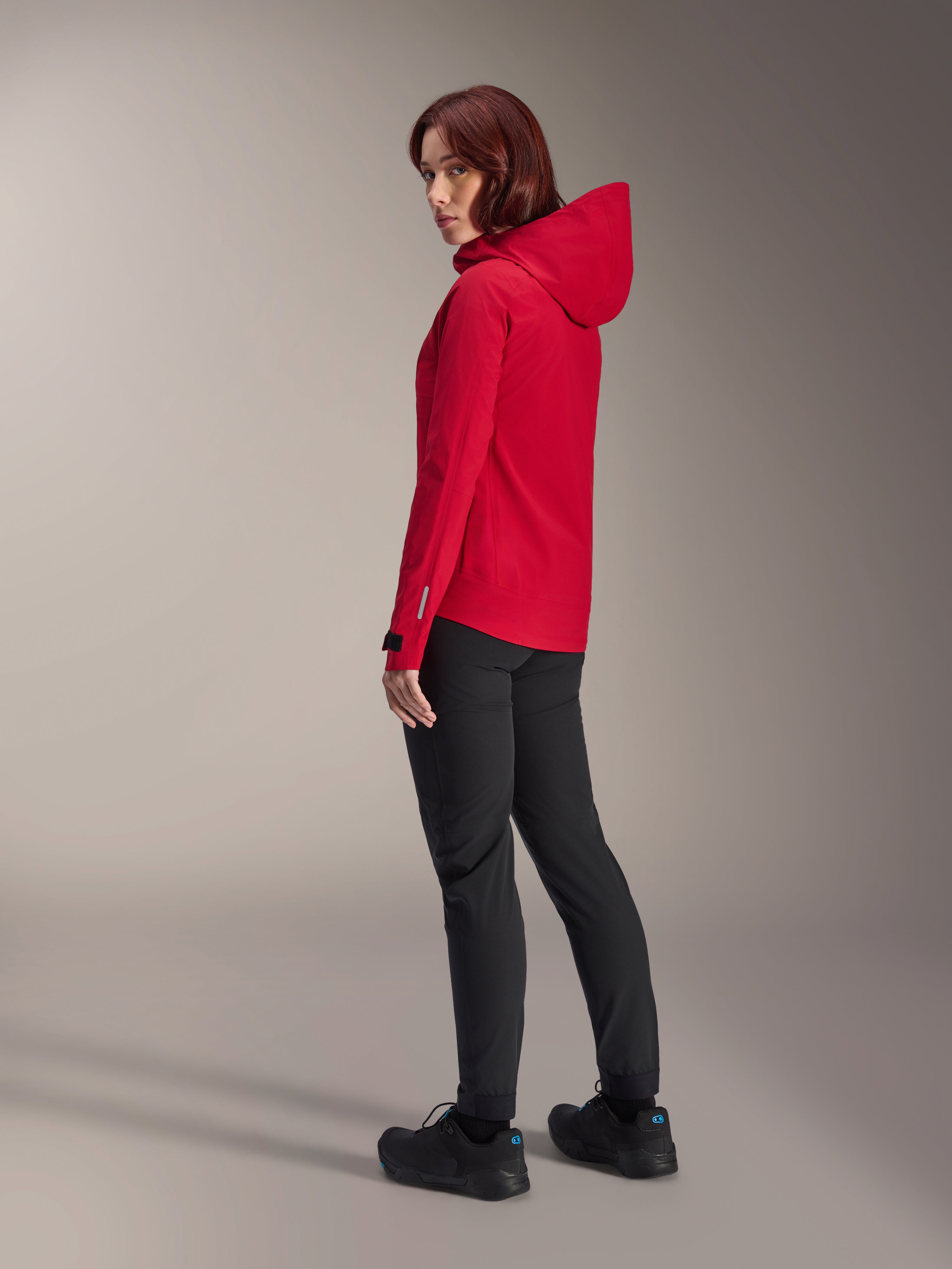 Mujer vistiendo la chaqueta impermeable Alpinestars Stella A-Dura Elite en color Red Fluo, combinada con pantalones técnicos de ciclismo negros y calzado negro, vista trasera de tres cuartos que muestra la capucha ajustable y el dobladillo trasero más largo, ropa de alto rendimiento para bicicleta de montaña.