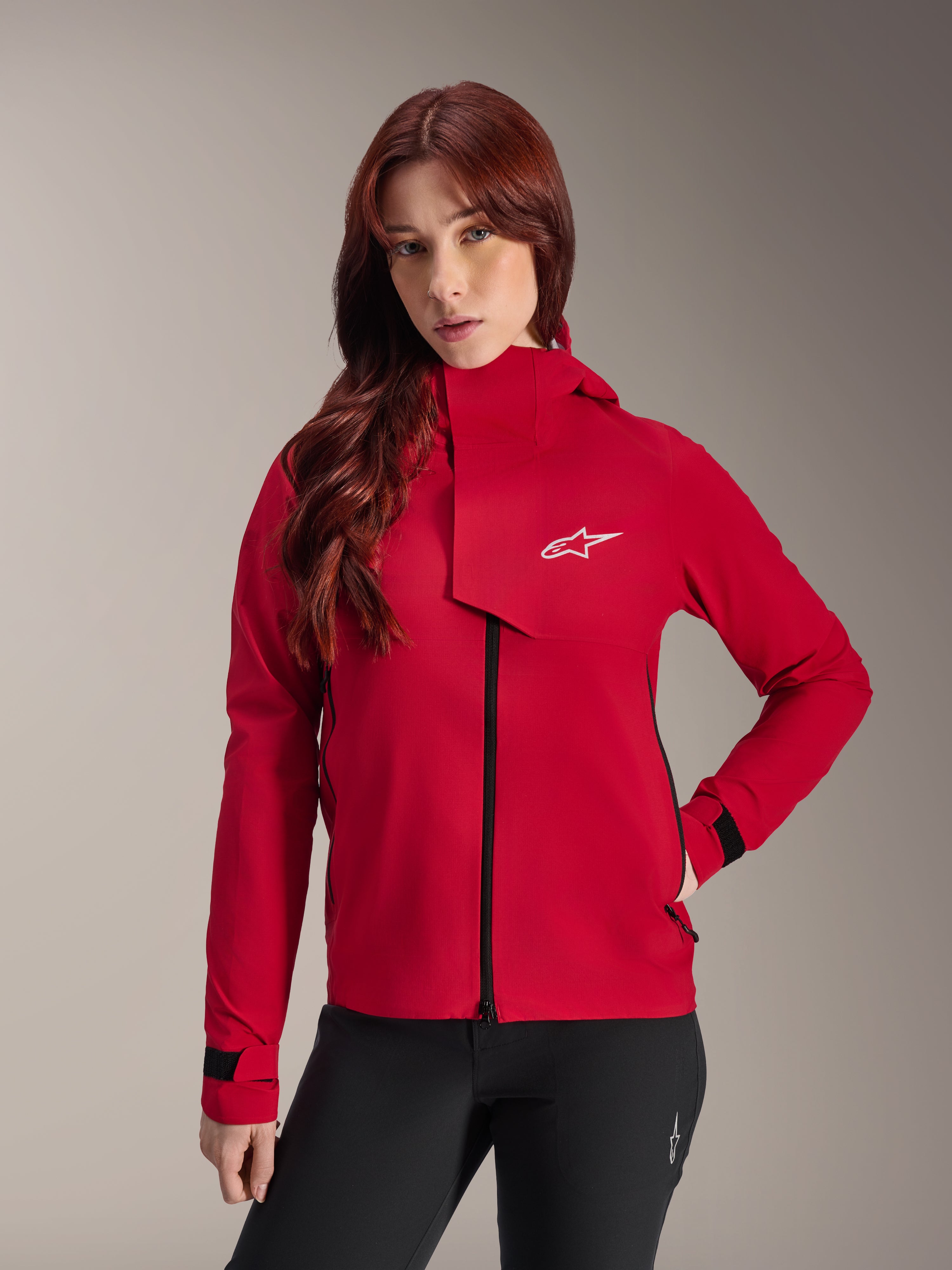 Chaqueta impermeable Alpinestars Stella A-Dura Elite, chaqueta de ciclismo de montaña para mujer, rojo flúor, con solapa frontal con el logotipo blanco de Alpinestars, cuello alto y puños ajustables, combinada con pantalones negros para un estilo de conducción técnico