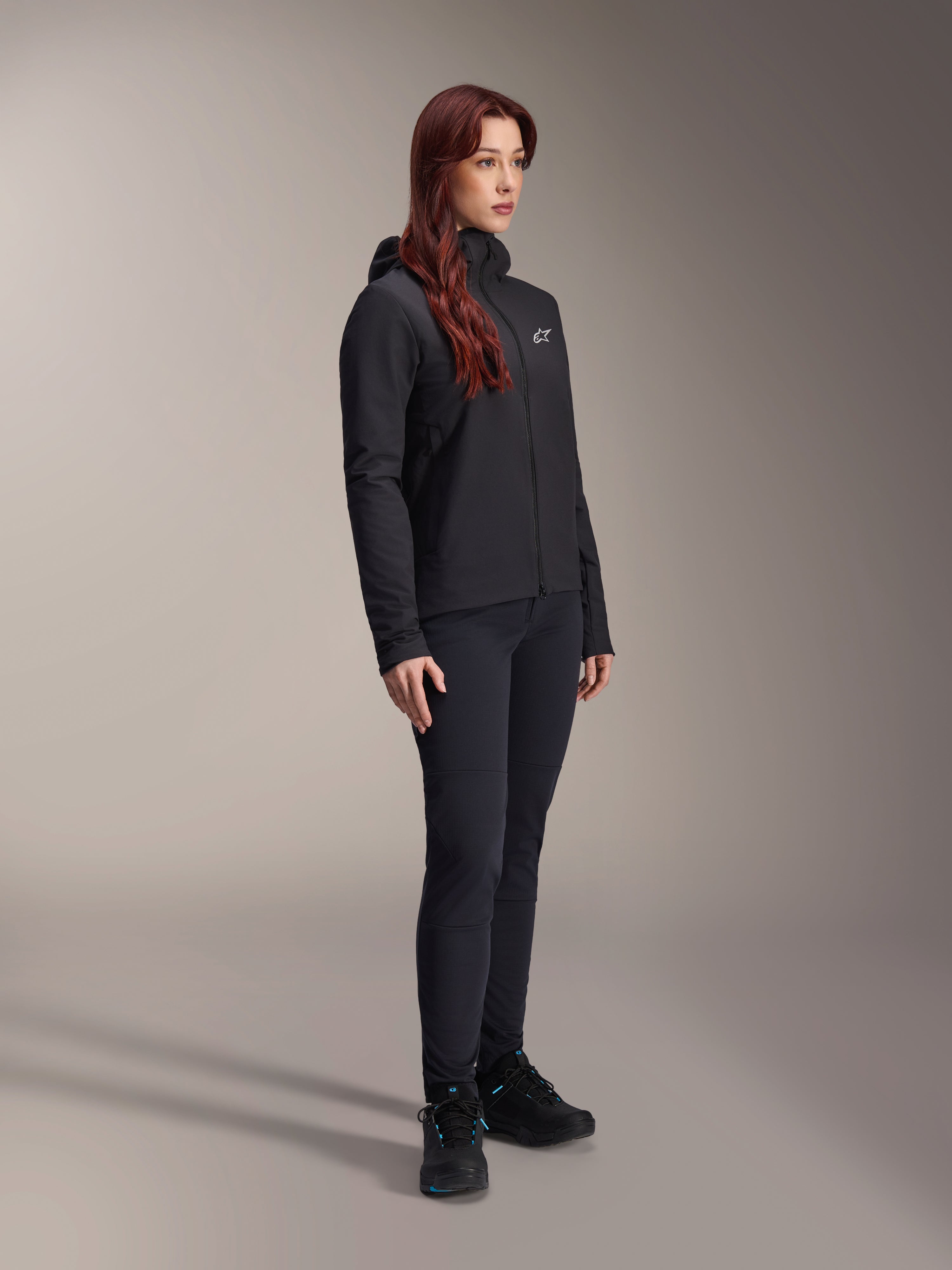 Alpinestars Stella A-Dura Thermal Jacket, chaqueta aislante ligera para mujer en color negro con capucha, combinada con pantalones negros y calzado negro, equipación profesional de ciclismo y MTB para condiciones de frío