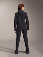 Mujer vistiendo la chaqueta térmica Alpinestars Stella A-Dura, vista trasera, chaqueta de ciclismo aislante negra con capucha, pantalones y calzado negros a juego, con un pequeño logotipo blanco en la parte inferior de la espalda