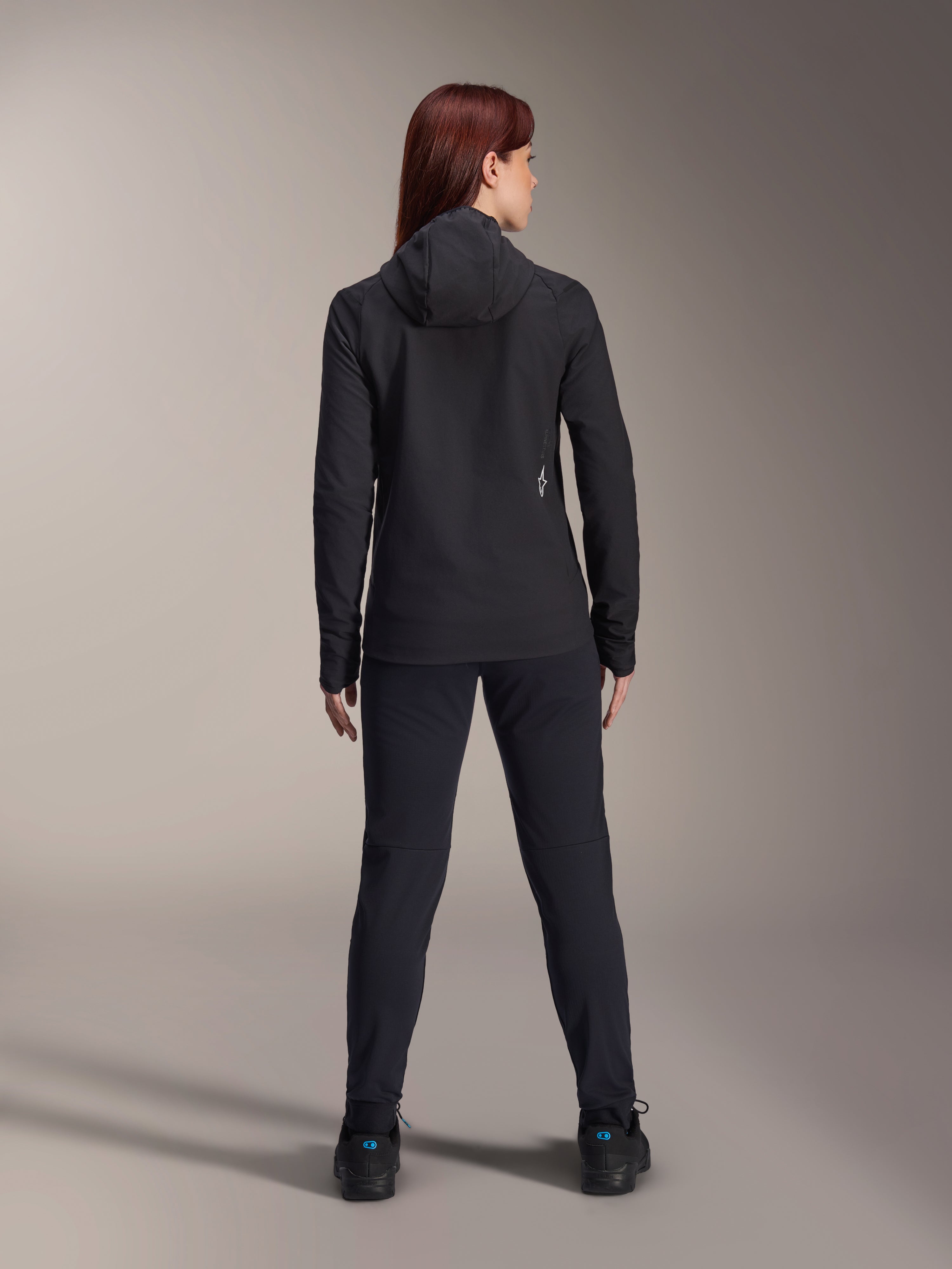 Mujer vistiendo la chaqueta térmica Alpinestars Stella A-Dura, vista trasera, chaqueta de ciclismo aislante negra con capucha, pantalones y calzado negros a juego, con un pequeño logotipo blanco en la parte inferior de la espalda