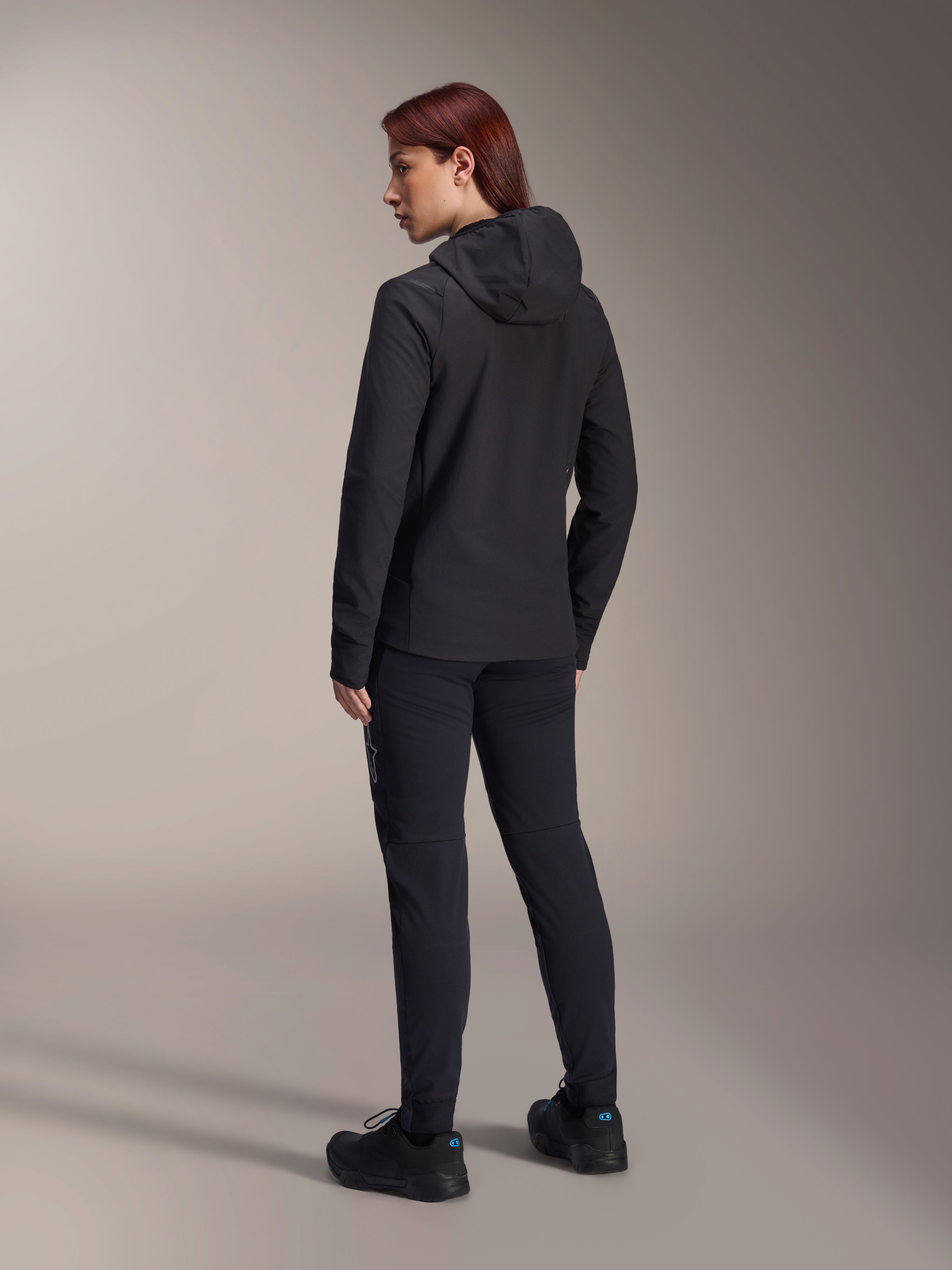 Chaqueta térmica Alpinestars Stella A-Dura, chaqueta de ciclismo MTB para mujer, negra, vista posterior que destaca la capucha aislante y el ajuste regular, combinada con pantalones y calzado negros a juego, aislamiento térmico ligero para rodar en climas fríos
