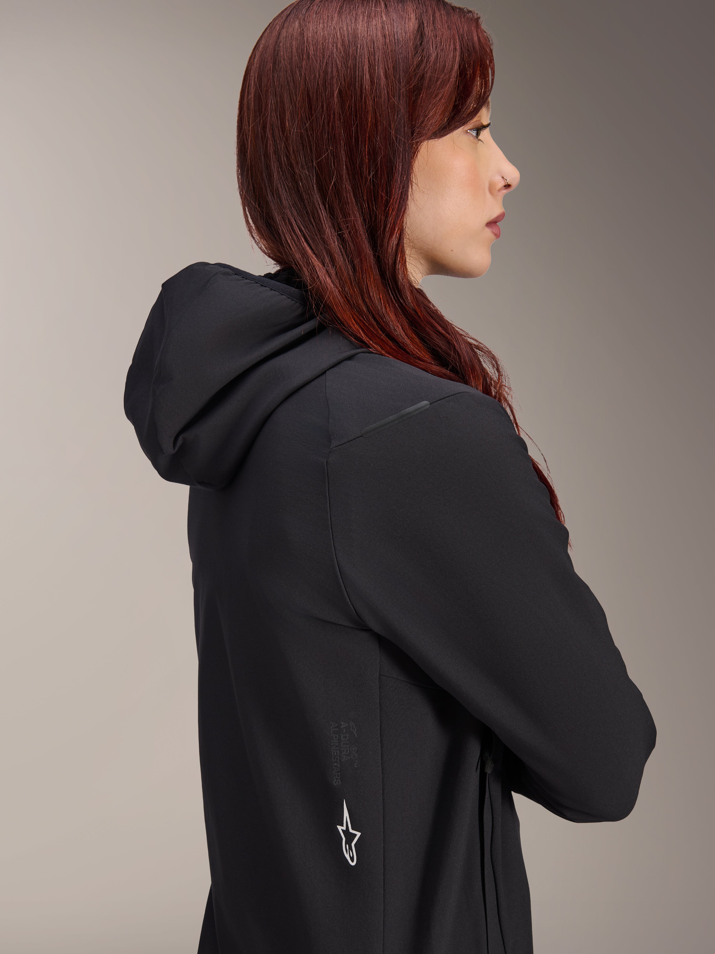 Chaqueta térmica Alpinestars Stella A-Dura, chaqueta de ciclismo para mujer, color negro, perfil lateral de una mujer con cabello pelirrojo luciendo la chaqueta, cuenta con capucha integrada y logotipo de la estrella de Alpinestars en blanco en el lateral, equipo térmico ligero y aislado para MTB
