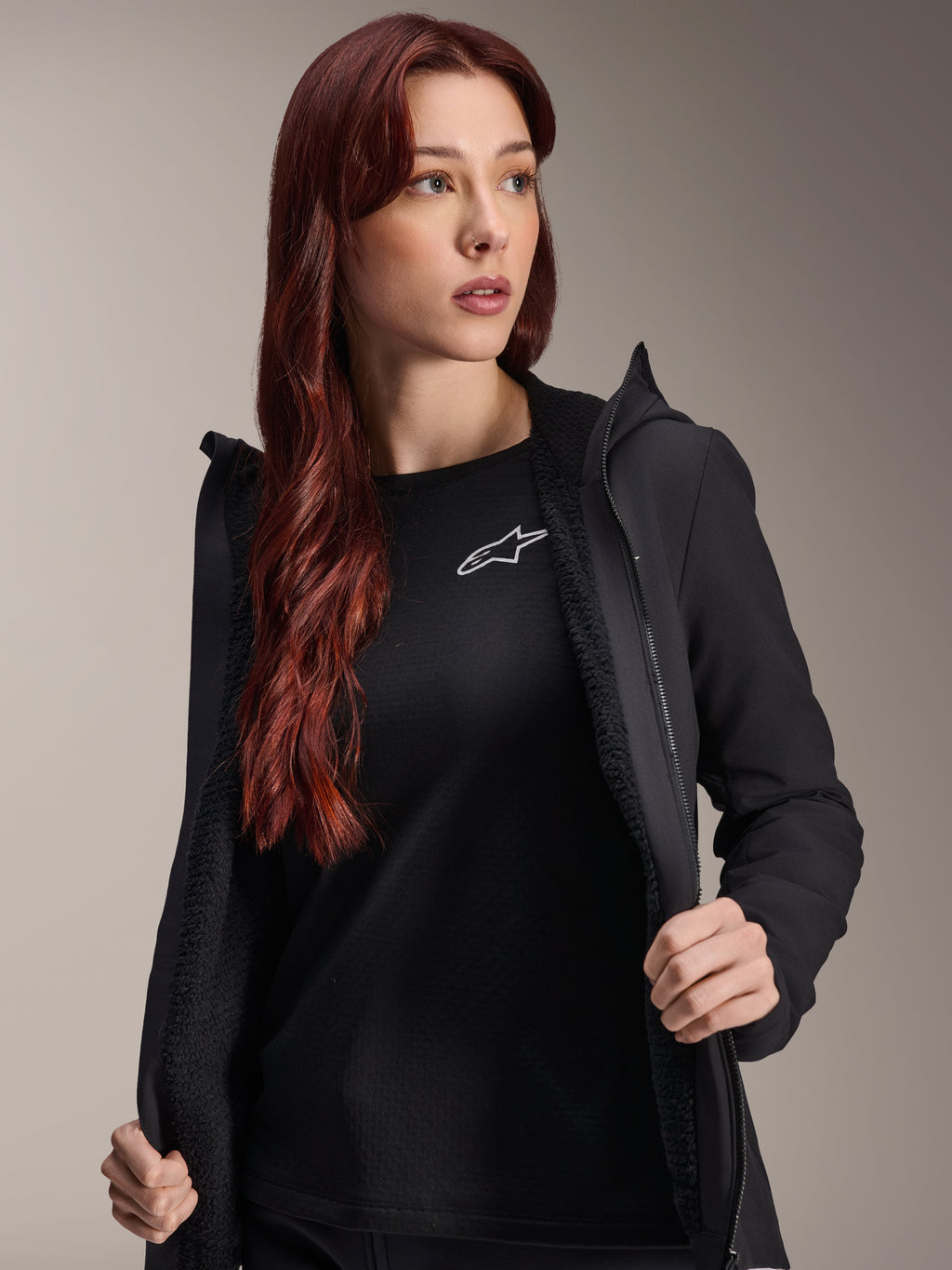 Alpinestars Stella A-Dura Thermal Jacket, chaqueta de ciclismo para mujer en color negro, mostrada abierta con forro de vellón texturizado y cremallera frontal, usada sobre una camiseta negra con el logotipo de Alpinestars, ligera y aislada para montar en climas fríos