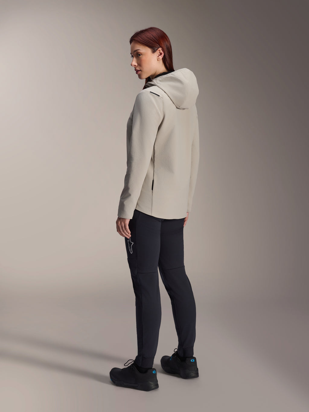 Chaqueta térmica Alpinestars Stella A-Dura en color gris titanio, vista trasera de tres cuartos de una mujer con la chaqueta de ciclismo con capucha y aislamiento y pantalones de MTB negros, toma de estudio que muestra el ajuste regular y el detalle del bolsillo.