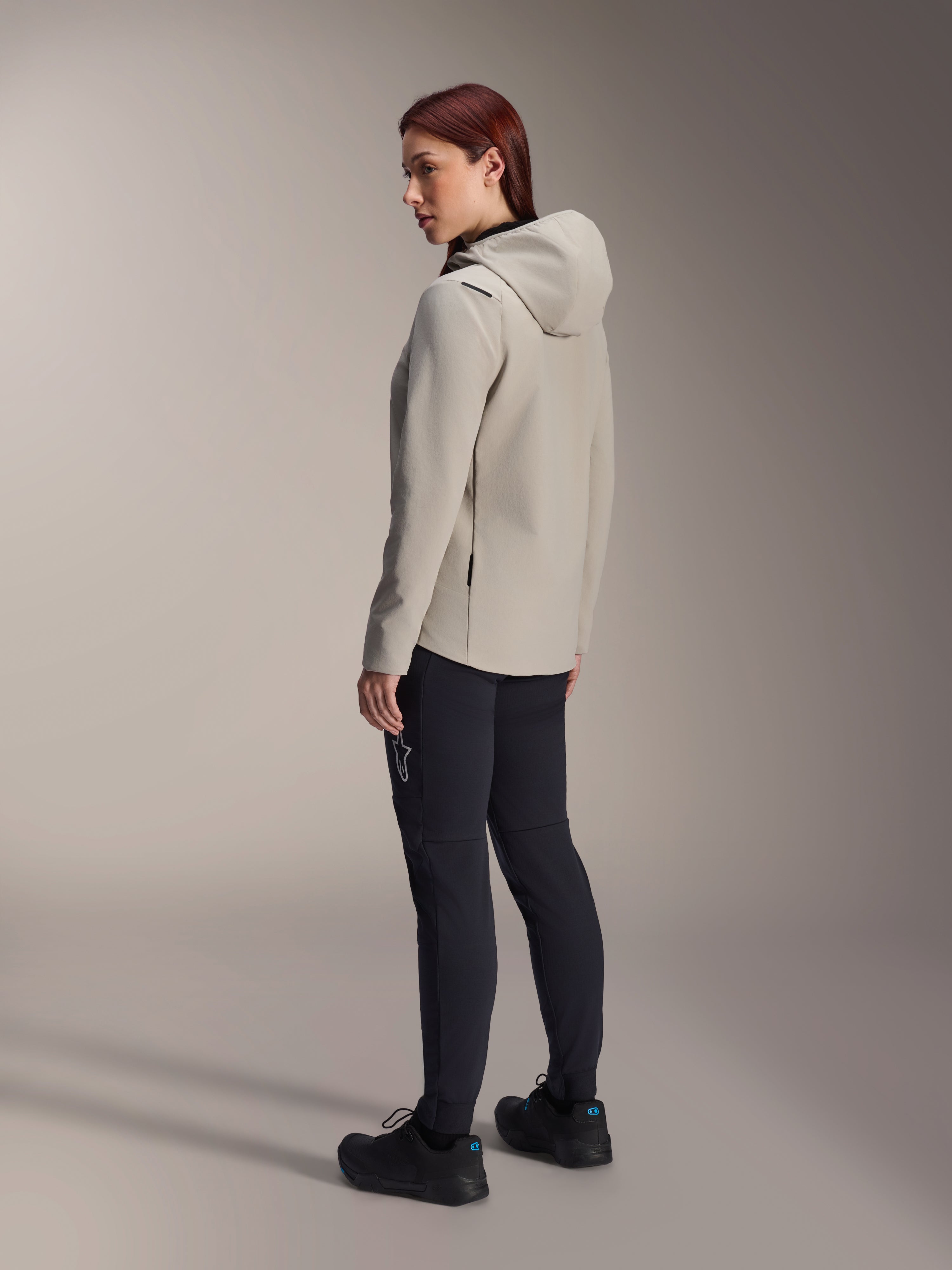 Chaqueta térmica Alpinestars Stella A-Dura en color gris titanio, vista trasera de tres cuartos de una mujer con la chaqueta de ciclismo con capucha y aislamiento y pantalones de MTB negros, toma de estudio que muestra el ajuste regular y el detalle del bolsillo.