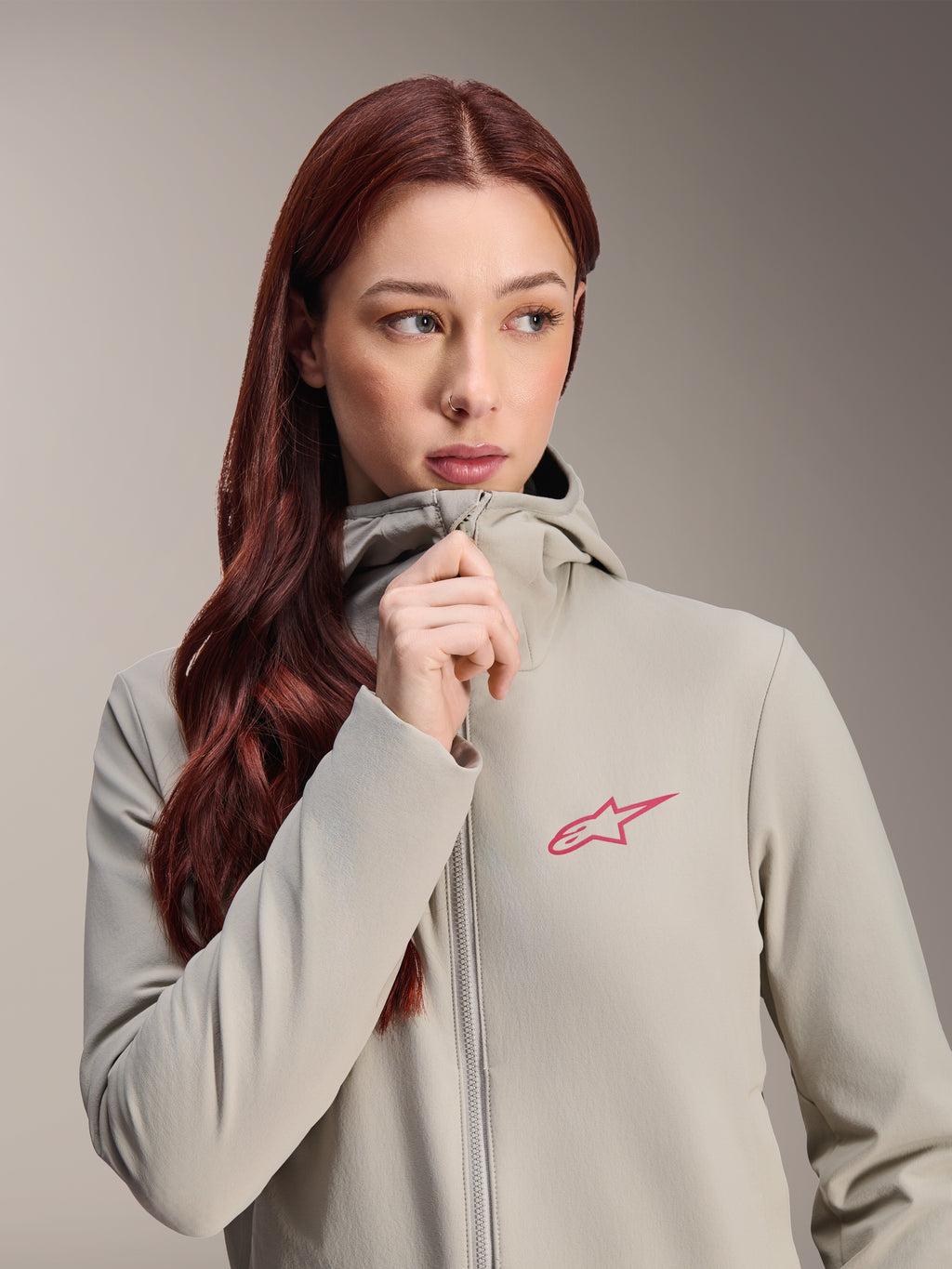 Chaqueta térmica Stella A-Dura de Alpinestars para mujer, gris titanio con un logotipo de estrella de Alpinestars en rosa, modelo con cabello largo y pelirrojo subiendo la cremallera de la chaqueta con capucha, ropa de ciclismo ligera con aislamiento para climas fríos