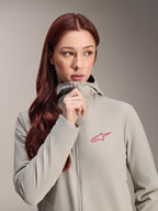 Chaqueta térmica Stella A-Dura de Alpinestars para mujer, gris titanio con un logotipo de estrella de Alpinestars en rosa, modelo con cabello largo y pelirrojo subiendo la cremallera de la chaqueta con capucha, ropa de ciclismo ligera con aislamiento para climas fríos