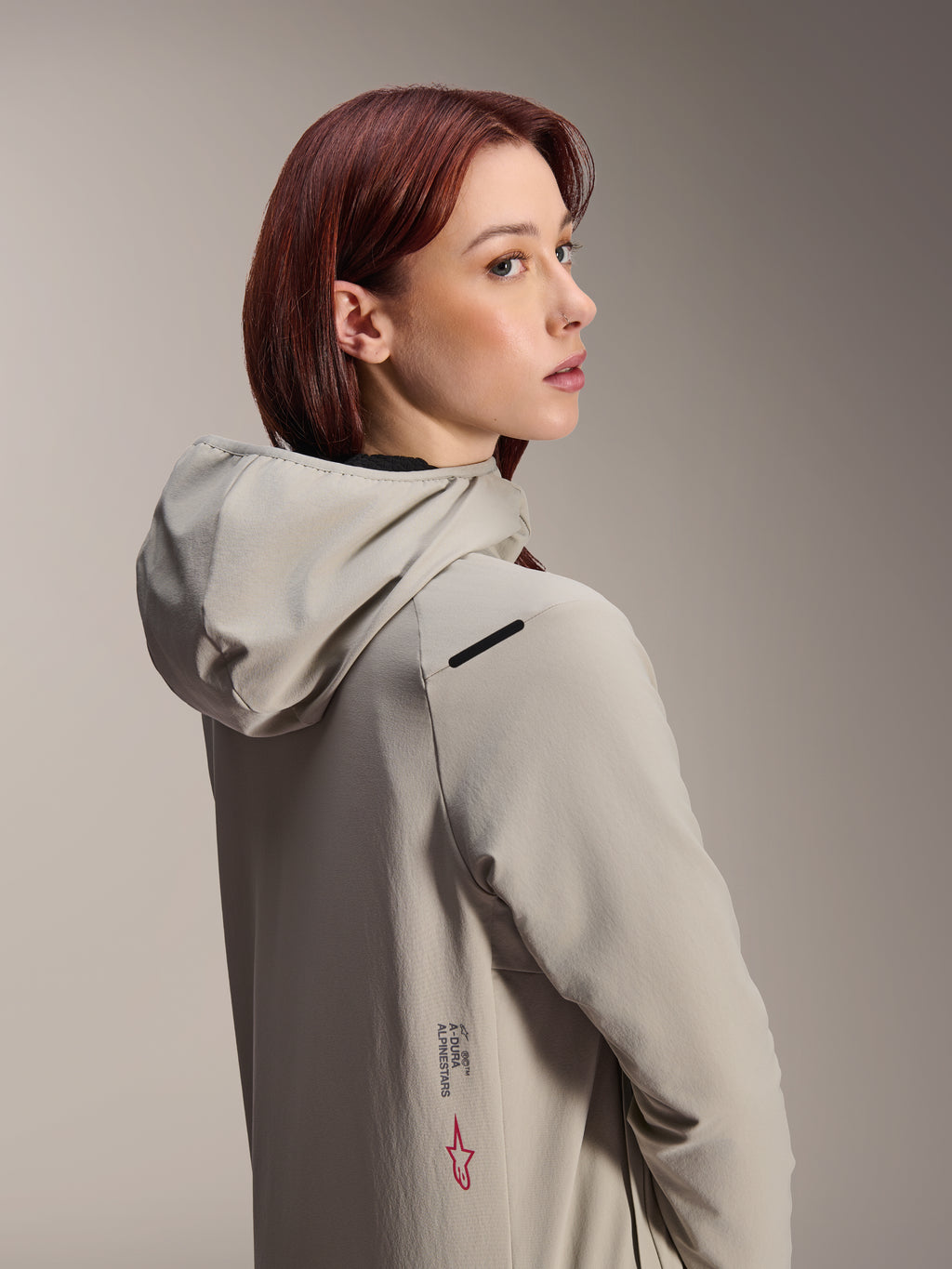 Alpinestars Stella A-Dura Thermal Jacket, chaqueta de MTB para mujer, gris titanio, mujer mirando por encima de su hombro con chaqueta técnica con capucha, con el texto Alpinestars y el logotipo de la estrella roja en el lateral, diseñada para ciclismo en climas fríos