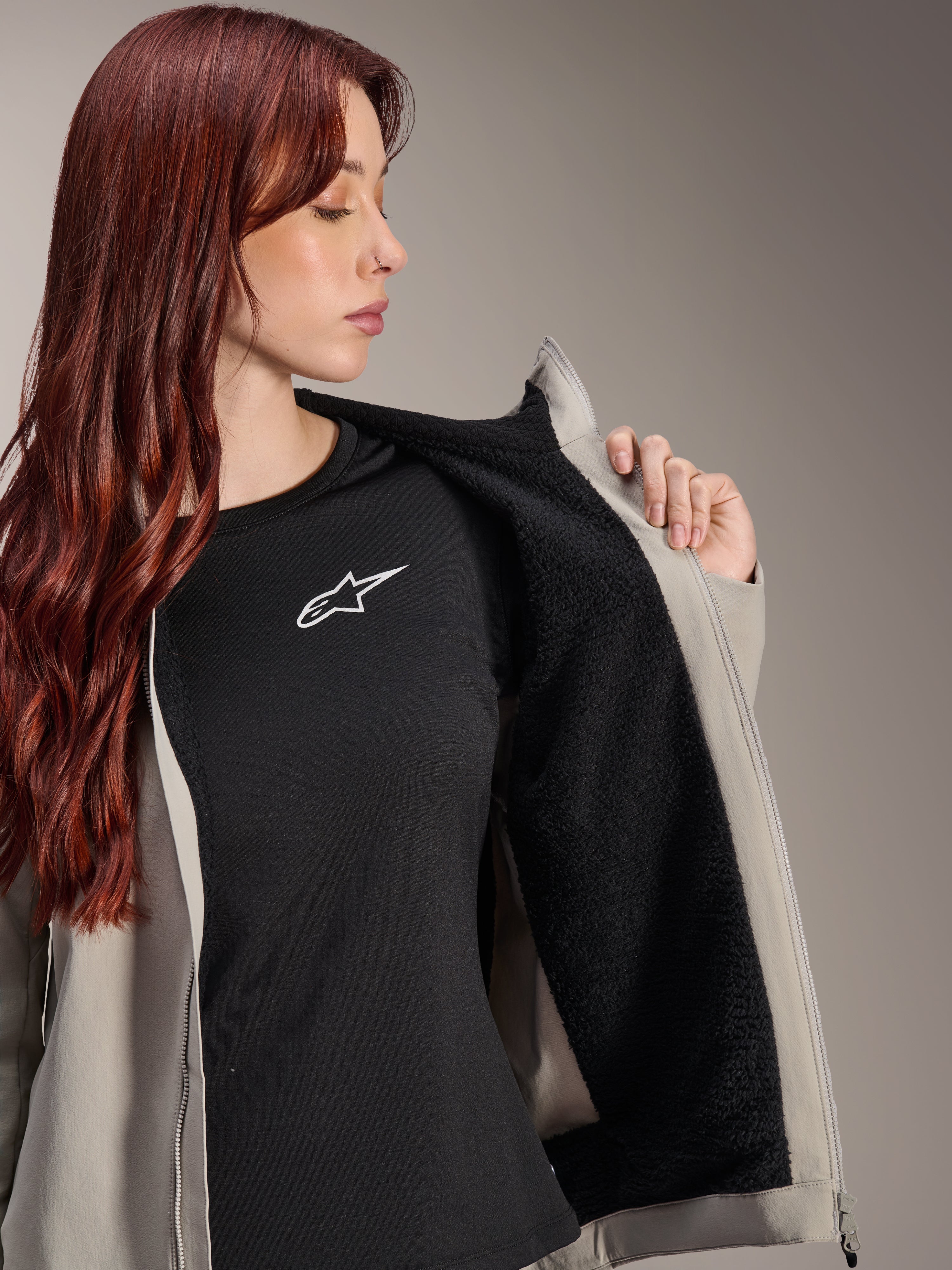 Mujer con la chaqueta térmica Alpinestars Stella A-Dura en Titanium, mostrando el forro de vellón negro texturizado, sobre una camiseta negra con logo de estrella blanca, chaqueta ligera de ciclismo de montaña para condiciones de frío