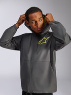 Chaqueta cortavientos Alpinestars Speeder Chromium, gris carbón y amarillo de alta visibilidad, modelo masculino con cortavientos con capucha y cordón, logotipo de Alpinestars en amarillo de alta visibilidad en el pecho y forro de la capucha en amarillo, ropa urbana casual inspirada en el motor