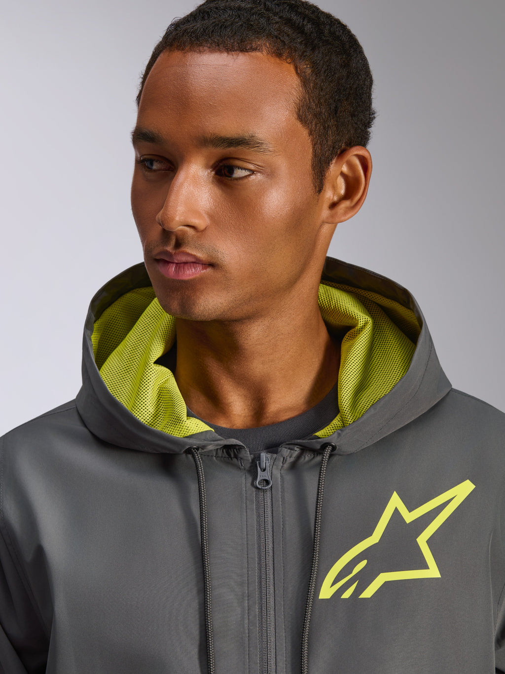 Chaqueta cortavientos Alpinestars Speeder Chromium, gris carbón con detalles en amarillo de alta visibilidad, con un logotipo de estrella amarilla en el pecho y forro de malla en la capucha a juego, cortavientos casual ligero