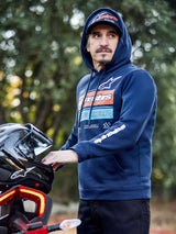 Sudadera Alpinestars Harken Hoodie, azul marino, sudadera de forro polar con gráfico degradado en el pecho y logotipo Alpinestars blanco en la manga, vestida por un hombre sujetando un casco de moto, ropa urbana casual inspirada en el motociclismo