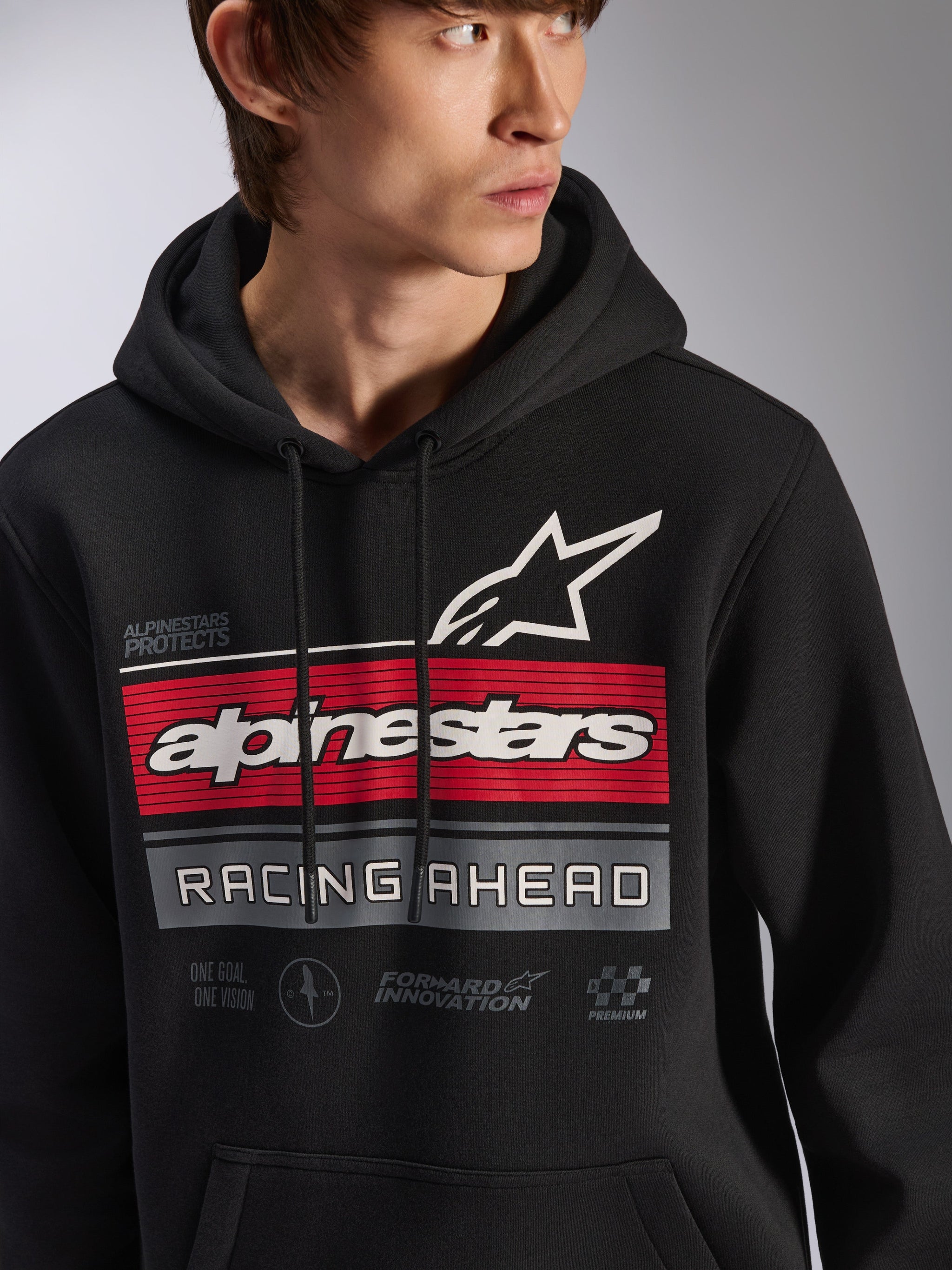 Sudadera con capucha Alpinestars Harken, sudadera negra de forro polar tipo pullover llevada por un modelo, con un gran gráfico en el pecho con franjas rojas y grises, el logotipo de la estrella y el nombre de Alpinestars en blanco, texto 'Racing Ahead', estilo urbano informal inspirado en el automovilismo moderno.
