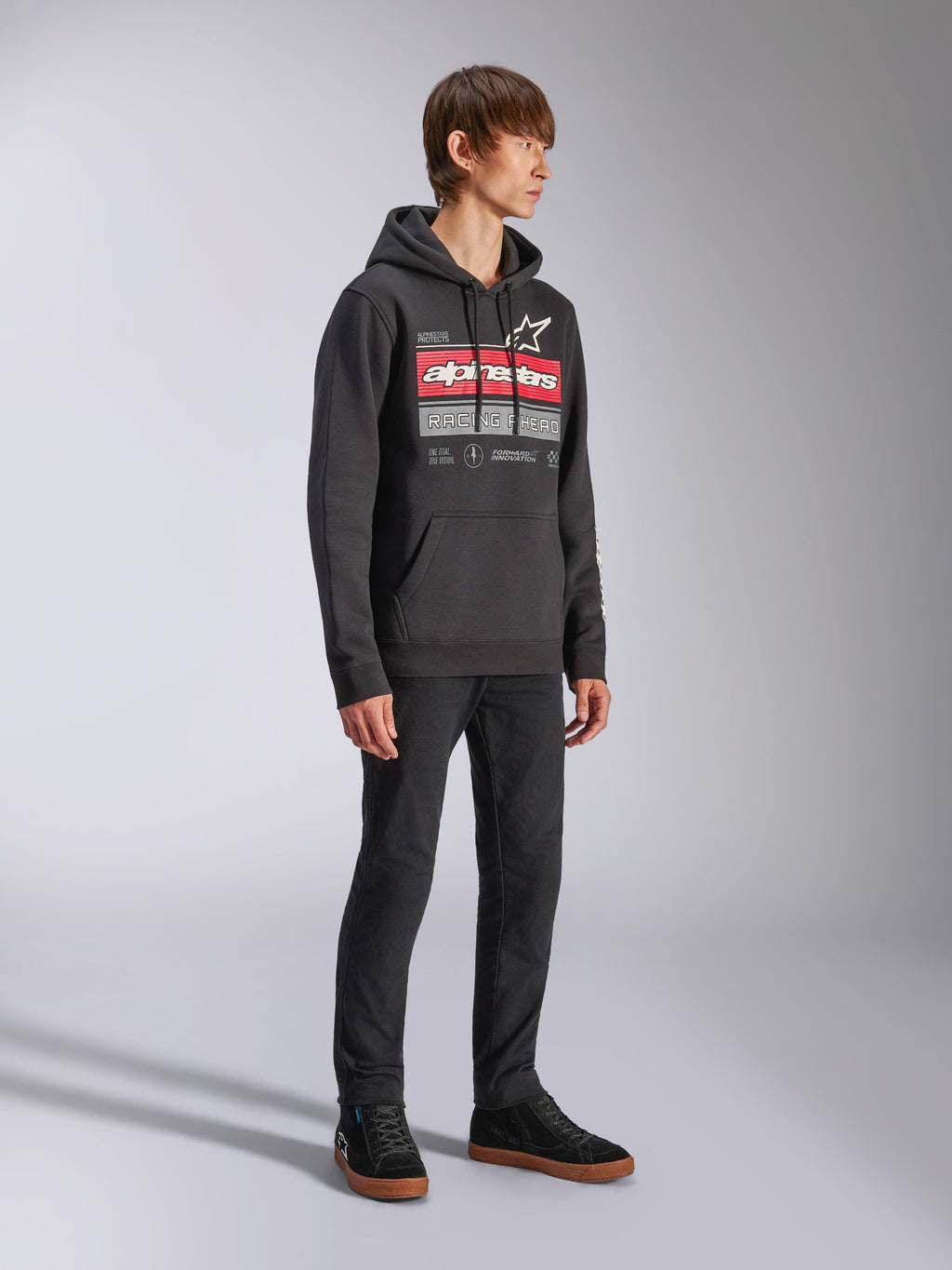 Sudadera con capucha Alpinestars Harken, sudadera negra tipo pullover de forro polar, con un gráfico grande en el pecho que destaca la marca Alpinestars en rojo y el logo de la estrella, incluye bolsillo canguro, vestida por un modelo con pantalones negros y zapatillas, estilo casual motorsport