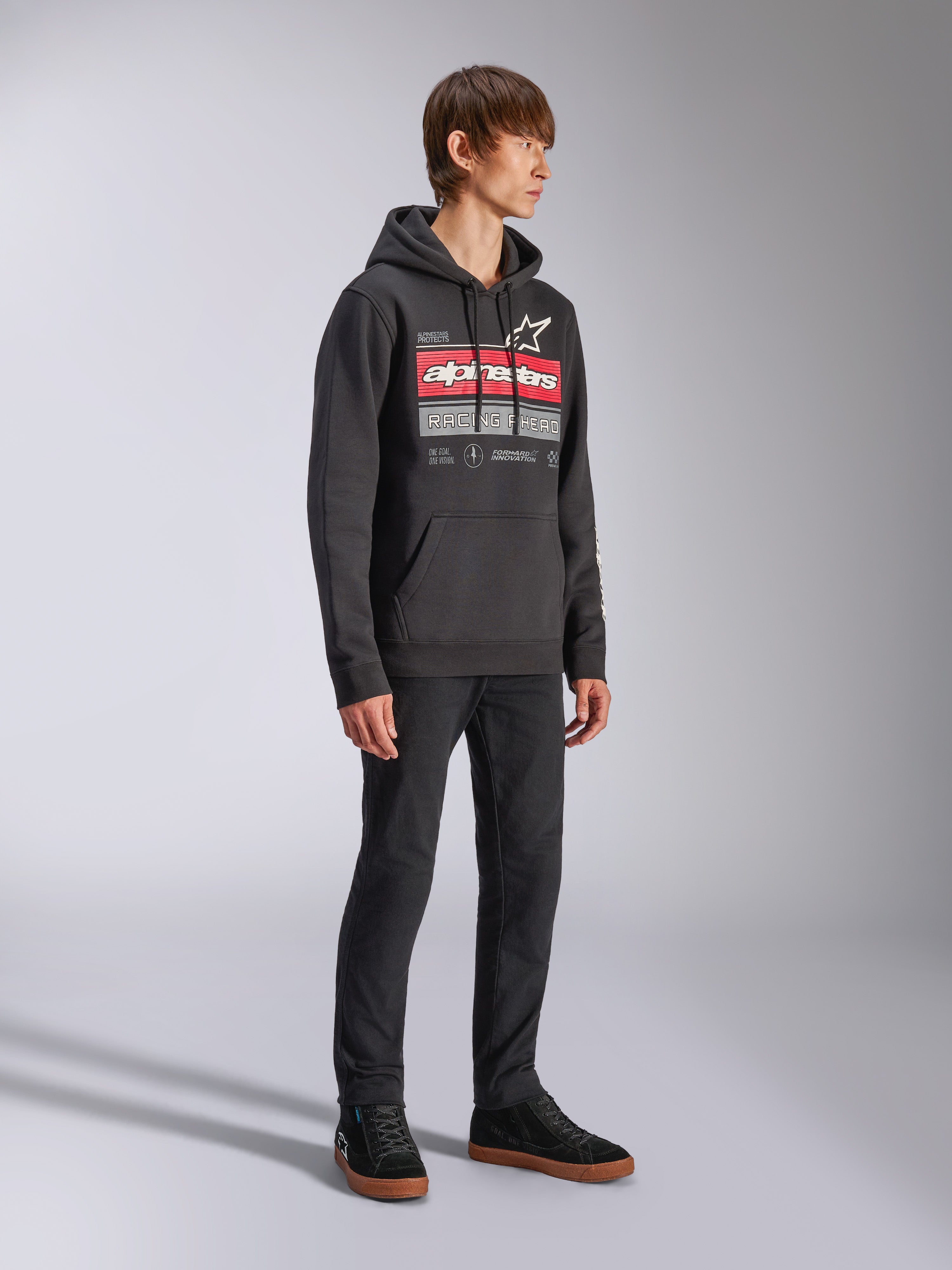 Sudadera con capucha Alpinestars Harken, sudadera negra tipo pullover de forro polar, con un gráfico grande en el pecho que destaca la marca Alpinestars en rojo y el logo de la estrella, incluye bolsillo canguro, vestida por un modelo con pantalones negros y zapatillas, estilo casual motorsport