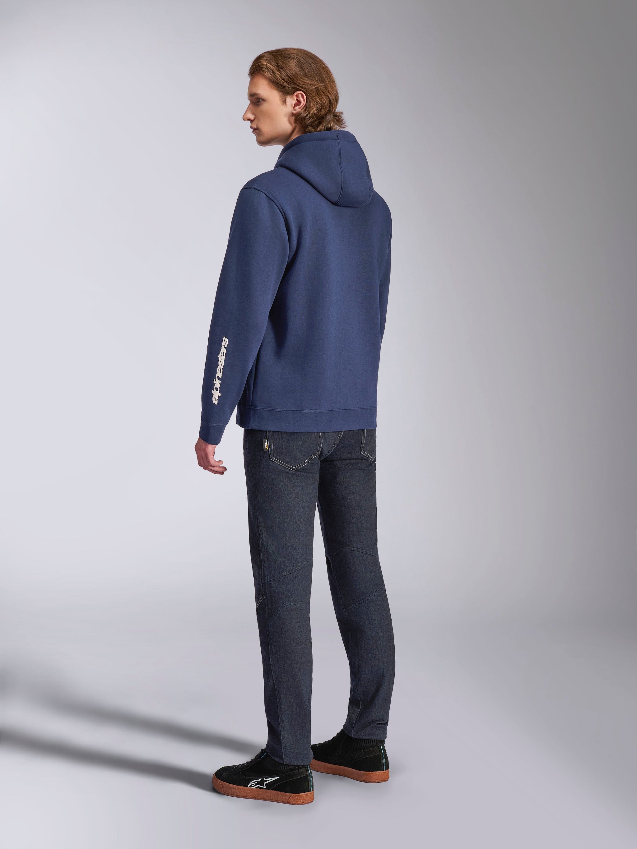 Sudadera con capucha Alpinestars Harken en azul marino, vista posterior de modelo masculino, sudadera informal de forro polar con logotipo de la marca en blanco en la manga, combinada con vaqueros de denim oscuro y zapatillas negras, ropa urbana moderna inspirada en el automovilismo