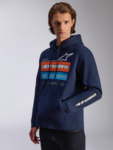 Alpinestars Harken Hoodie, azul marino, llevada por un modelo, con un gráfico llamativo en el pecho con diseño 'Racing Ahead' en naranja y azul, logotipo Alpinestars en la manga, bolsillo canguro y capucha con cordón